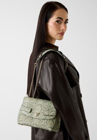 Torebka o wzorze zielonej skóry węża z złotymi okuciami i łańcuszkowym paskiem, z klapą i trójkątnym logo na przedniej stronie.