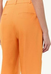 Orangefarbene Hosen mit glatter Stofftextur, ausgestatteten Gesäßtaschen und taillierter Passform. Abgeschlossen mit dezenten Nahtdetails.