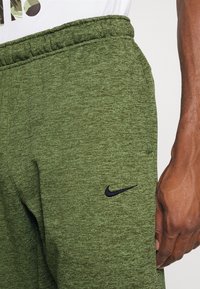 Calças de fato verde mescla feitas de tecido suave, com um logótipo da Nike em preto, cintura elástica e bolsos laterais. Vista de perto da textura.