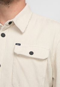 Beige Corduroy-Hemd mit spitzem Kragen, ausgestattet mit schwarzen Druckknöpfen und einer Brusttasche mit einem Logosticker. Vertikale Rippstruktur.