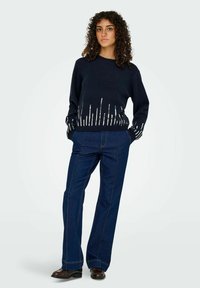 Marineblauw gebreid sweater met wit verticale streep patroon aan de zoom, gecombineerd met blauwe wijd uitlopende denim broeken en donkere schoenen.