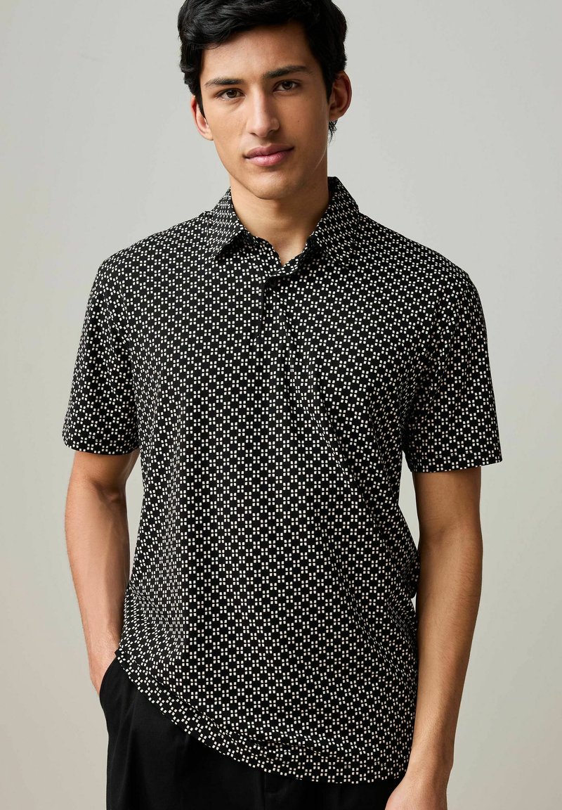 Camicia a maniche corte, con colletto, di colore nero con motivi geometrici bianchi. Realizzata in materiale leggero, presenta una tasca sul petto.
