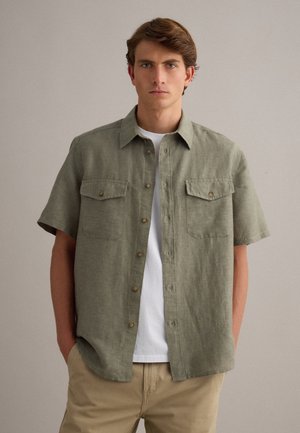 Junger Mann mit kurzen braunen Haaren, der ein olivgrünes kurzärmeliges Hemd über einem weißen T-Shirt und beigen Hosen trägt, steht mit den Händen in den Taschen.