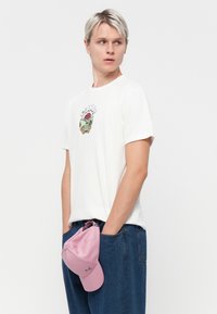 White short-sleeve t-shirt featuring a colorful graphic print on the front, paired with blue denim trousers and a pink cap held in the pocket.
