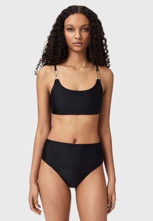 Slank kvinde med krøllet hår iført en sort højtaljet bikini med guldspændebånd, stående mod en ensfarvet lys baggrund.