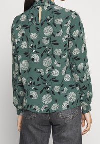 Green floral blouse featuring white and black patterns, long sleeves with gathered cuffs, high neckline with button closure, and keyhole detail.