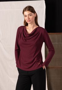 Burgundy long-sleeve top with draped neckline, made of soft fabric, paired with black trousers. Features subtle gold branding detail.