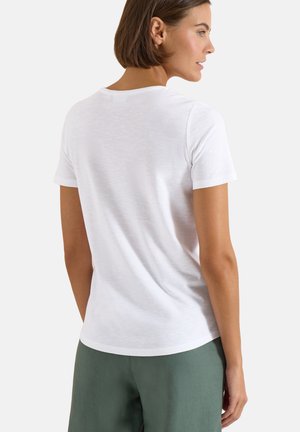 Vrouw met kort bruin haar, die een eenvoudig wit T-shirt met korte mouwen en een groene broek draagt, van achteren en in zijaanzicht.