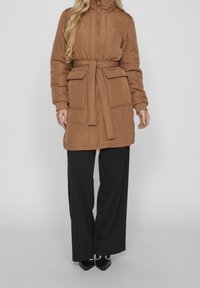 Manteau matelassé marron avec un col montant, une taille cintrée et de grandes poches avant, associé à un pantalon large noir et des chaussures à bouts pointus.