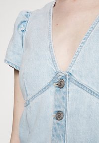 Vestido de mezclilla azul claro con escote en V, mangas cortas abullonadas y dos botones metálicos en contraste en la parte frontal con detalles de costura visibles.