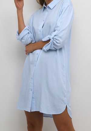 Femme portant une robe-chemise à manches longues bleu clair, avec les manches retroussées, debout devant un fond clair uni.