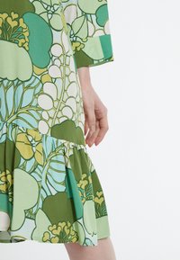 Blommig klänning med en grön och ljusblå färgpalett, strukturerad byst och rynkad volangkjol. Texturdetalj synlig i tyget.