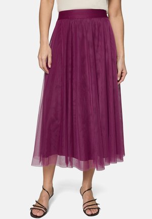 Femme portant une jupe midi en tulle violet avec une taille ajustée et des sandales noires à lanières, debout devant un fond blanc.
