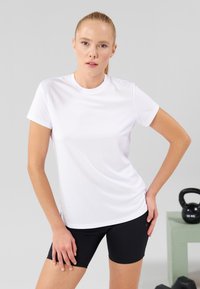 Tricou sportiv alb cu mâneci scurte și gât rotund, asortat cu pantaloni șort negri. Material neted și design ajustat. O kettlebell în fundal.