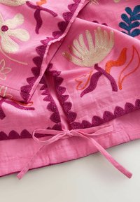 Pink fabric with embroidered flowers in cream, purple, orange, and blue, featuring scalloped edges and a tied bow at the bottom center.