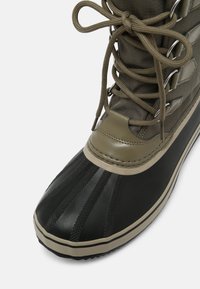 Bota impermeable con una puntera de goma negra, parte superior de tela verde oliva, ojales reforzados y cordones, con una superficie texturizada y un patrón en relieve.