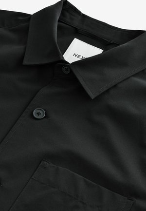 Camisa negra de botones con cuello, bolsillo frontal y una textura suave y mate. Etiqueta visible dentro del cuello. Dos botones oscuros.