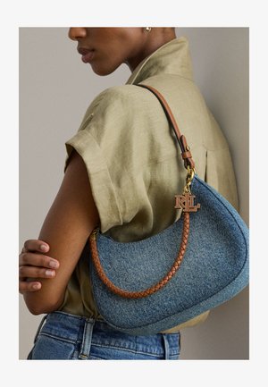 Femme en chemise beige aux manches retroussées tenant un sac à main en jean bleu avec une sangle en cuir tressé marron et un bijou doré RL sur l'épaule.