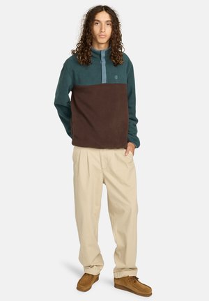 Fleece pullover med en turkis øverste sektion, brun nederste sektion, knaplukning ved halsen, samt afslappede beige bukser og brune sko.
