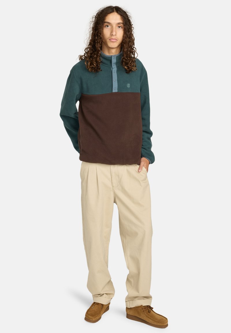 Fleece-pullover med en teal övre del, brun nedre del, knäppt halsringning, tillsammans med avslappnade beige byxor och bruna skor.