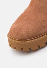 Suede ankle boot in tan with a thick, textured rubber sole featuring deep grooves for traction. Rounded toe design, clean stitching detail.