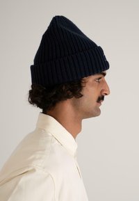 Navy knitted beanie with a ribbed texture, featuring a slouchy peak design. Worn with a light-colored collared shirt.
