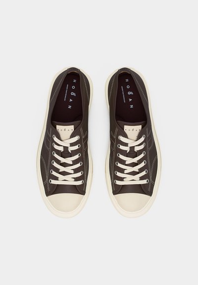 Par de zapatillas bajas de color marrón oscuro con suelas de goma blancas, punteras crema, cordones blancos y marca visible "Hogan" en las plantillas y lengüetas.