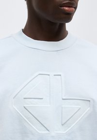 Κοντινό πλάνο ενός ατόμου που φοράει ανοιχτό μπλε φούτερ με μεγάλο ανάγλυφο γεωμετρικό λογότυπο στο στήθος.