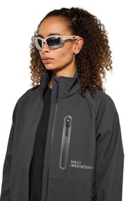 Graue Softshelljacke mit hohem Kragen, einer mit Reißverschluss versehenen Brusttasche und bedruckten Textdetails. Das Modell trägt eine Sonnenbrille mit weißen Rahmen.