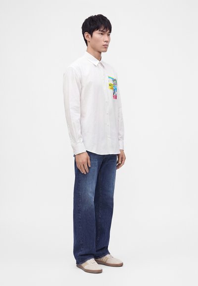 Homme portant une chemise blanche à boutons avec des patchs colorés, un jean bleu et des baskets blanches, debout devant un fond blanc uni.