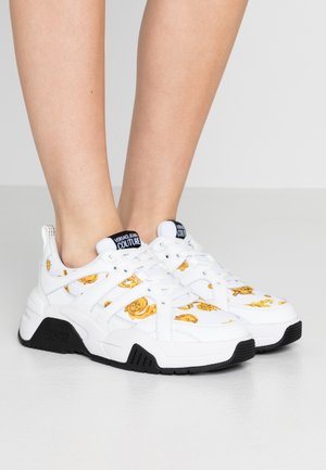 Białe sneakersy Versace Jeans Couture z złotym barokowym wzorem i czarną podeszwą, noszone na stopach na jednolitym tle.
