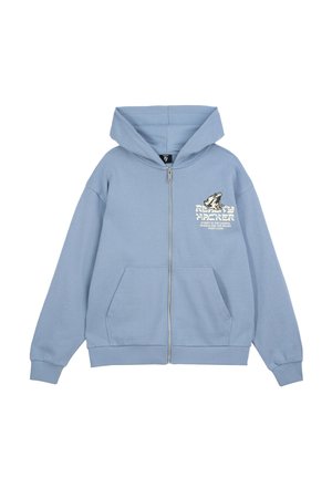 Młody mężczyzna stojący, ubrany w jasnoniebieską bluzę na zamek z grafiką, czarne, oversize spodenki do kolan, czarne trampki i białe skarpetki.