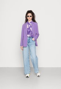 Blazer roxo sobre uma camisa com efeito tie-dye roxa e branca, combinada com jeans de perna larga azul clara e ténis brancos com detalhes amarelos.