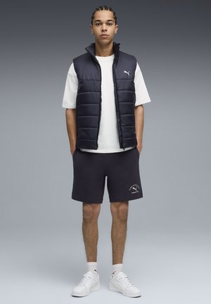 En marineblå puffervest over en hvid T-shirt, parret med mørke shorts med et logo. Hvide sneakers fuldender det atletiske look.