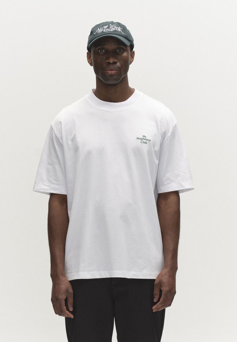 Homme portant un T-shirt blanc oversized avec le texte "The Neighbour Club" et une casquette de baseball sombre avec des lettres blanches, debout devant un fond uni.