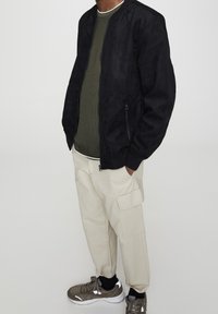 Blouson aviateur en daim noir, chemise côtelée olive, pantalon cargo beige avec poche latérale, baskets grises avec accents blancs. Design simple et moderne.