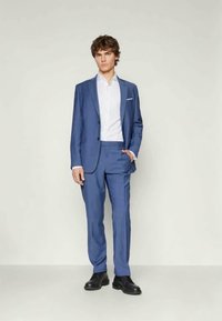 Blauer Anzug mit slim fit, einreihiger Jacke, Reverskragen und passender Hose. Weißes Hemd darunter und schwarze Schuhe.