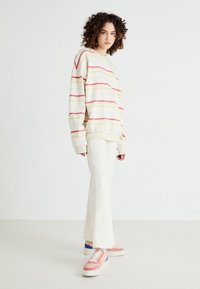 Pull uni crème à rayures roses, vertes et bleues ; coupe décontractée ; associé à un pantalon blanc évasé et des baskets roses avec des accents bleus.