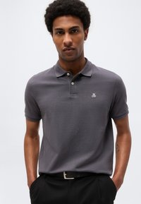 Νεαρός άνδρας φοράει γκρι πόλο πουκάμισο με λογότυπο "M OP", μαύρο παντελόνι και μαύρη ζώνη, στέκεται με τα χέρια στις τσέπες μπροστά σε απλό φόντο.