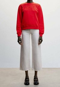 Sweatshirt rouge avec broderie détaillée au niveau du col, associé à un pantalon en denim blanc à jambes larges et des mocassins noirs avec des accents dorés.