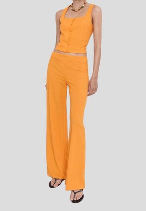 Mujer con top corto sin mangas de color naranja brillante con botones y pantalones de pierna ancha a juego, con sandalias negras y collar de cuentas.