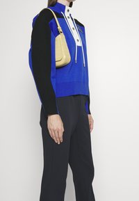 Kvinna som bär en blå och svart långärmad tröja med vita knappar och huva snören, svarta byxor och en beige axelväska.