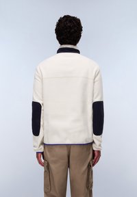 Fleece jacket in cream with dark blue collar and elbow patches. Features a straight cut, elastic cuffs, and a contrasting blue hem.