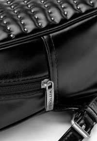 Bolso de cuero negro con textura acolchada, tachuelas plateadas, cierre con cremallera y tira con logo, además de una correa de hombro desmontable.