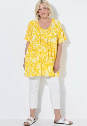 Kvinde stående iført en gul blomster tunika med korte ærmer, hvide kortere bukser og hvide åbne sandaler på en hvid baggrund.