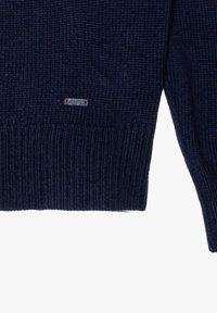 Maglione navy lavorato a maglia con polsini e orlo a coste. Presenta un piccolo etichetta metallica con dettagli rossi vicino al fondo. Texture morbida.