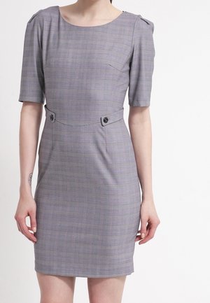 Kobieta ubrana w dopasowaną sukienkę w szarą kratę, z rękawami do łokci, okrągłym dekoltem i ozdobnymi guzikami w talii.