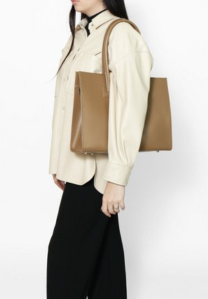 Borsa tote marrone chiaro, strutturata, con manici lunghi e lisci, caratterizzata da un design minimalista e una finitura elegante. Indossata sulla spalla.