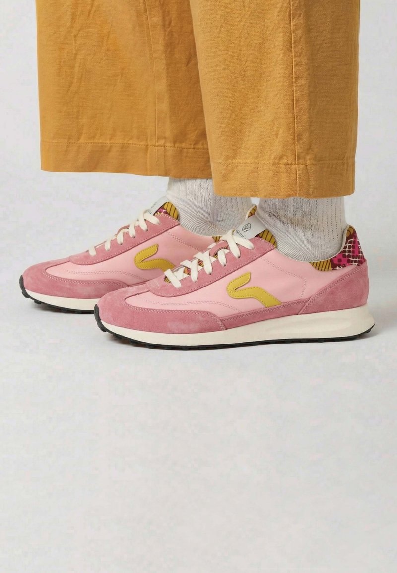 Personne portant des baskets roses avec des touches jaunes, des chaussettes blanches et un pantalon couleur moutarde, debout sur un sol clair.