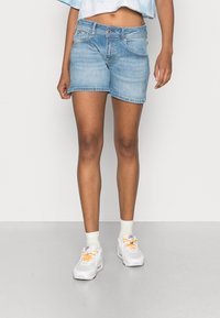 Denim shorts in light blue, featuring a classic five-pocket design, straight cut, and frayed hem. Paired with white sneakers and socks.